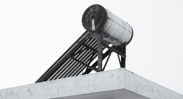 Metal cylindrical device on a rooftop, greyscale image aesthetic, light industrial rust visible, evacuated tube solar water heater in center of frame.
