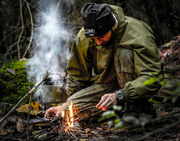 A man kneels in the forest, starting a small fire for cooking.